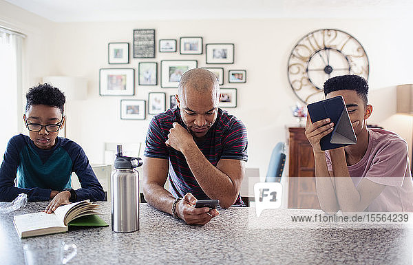 Vater und Söhne benutzen Tablet und Smartphone und lesen ein Buch in der Küche