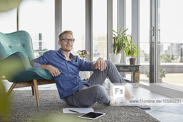 Lächelnder junger Mann sitzt auf Teppich mit Tablette und Notizblöcken zu Hause