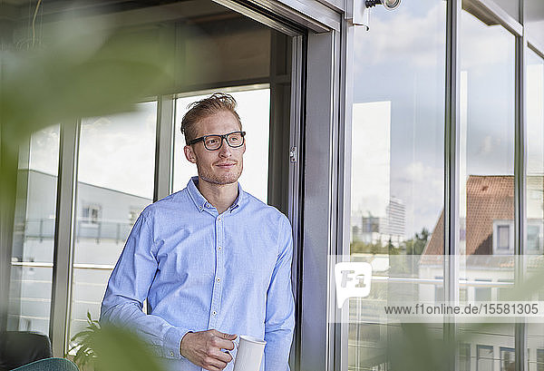 Young man with cup of coffee standing at balcony door