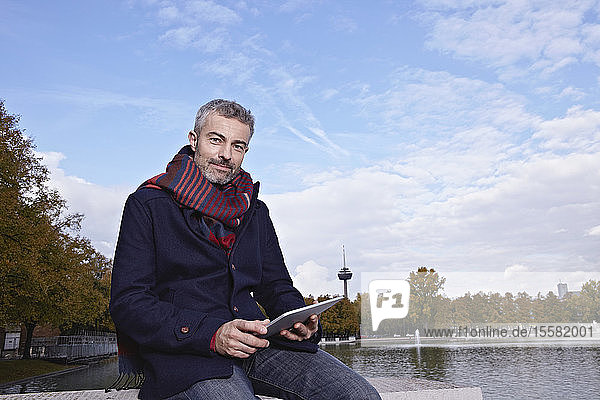 Deutschland  Köln  Erwachsener Mann sitzt auf Brücke mit digitalem Tablett  lächelnd  Porträt