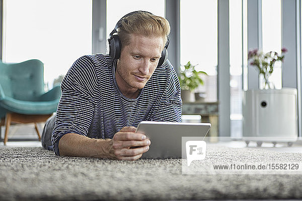 Young man lying on carpet at home wearing headphones and using tablet