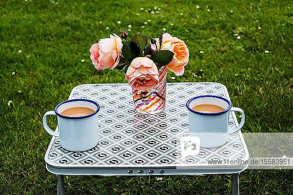Hochformatige Nahaufnahme von Tee in weißen Emaillebechern und Vase mit lachsrosa Rosen auf dem Picknicktisch.