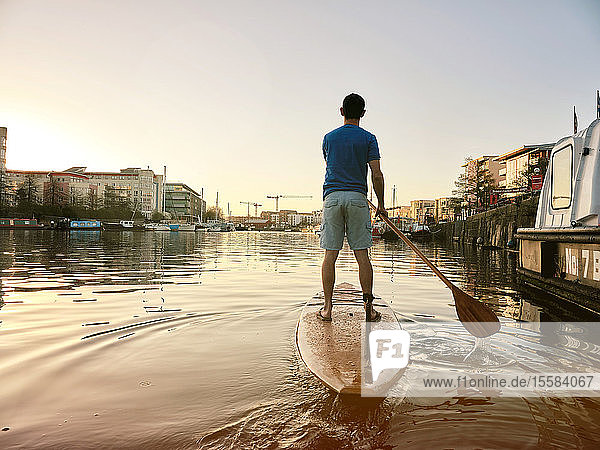Mann steht in der Morgendämmerung auf einem Paddelbrett auf einem Fluss  Schuss von hinten