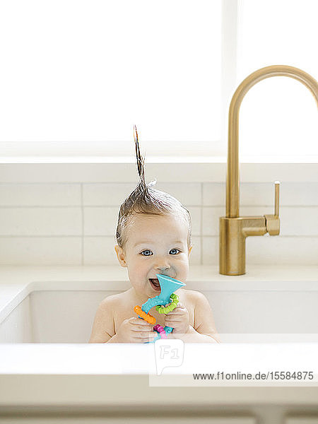 Baby girl with Mohican hairstyle holding toy while bathing in kitchen sink