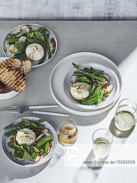 Stilleben in hoher Tonart mit Weißweingläsern und Tellern mit Burrata-Frühlingssalat auf weißem Tisch  Draufsicht