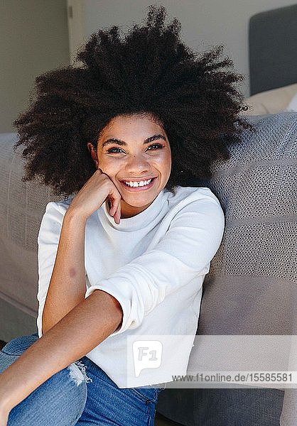 Fröhliche junge Frau mit Afrofrisur sitzt auf dem Schlafzimmerboden  Porträt