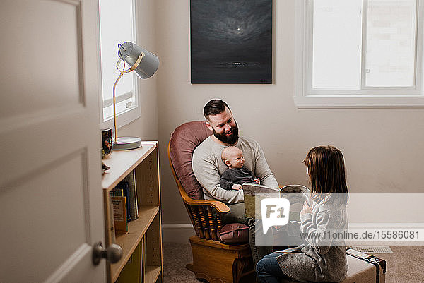 Girl in living room showing book to father and baby brother
