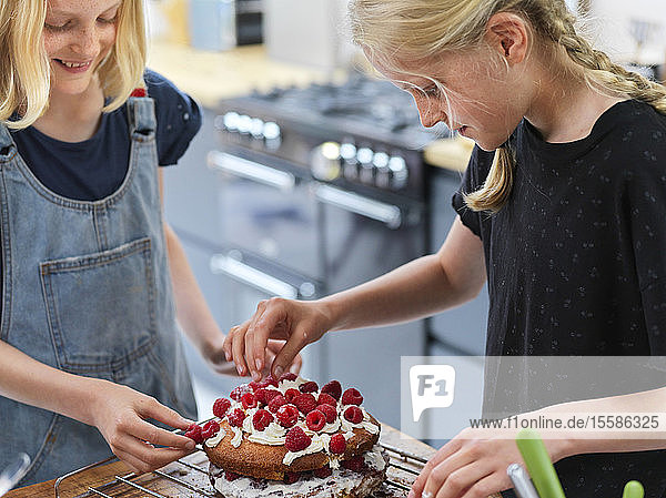 Girl and her sister baking a cake  decorating cake with fresh cream and raspberries at kitchen table