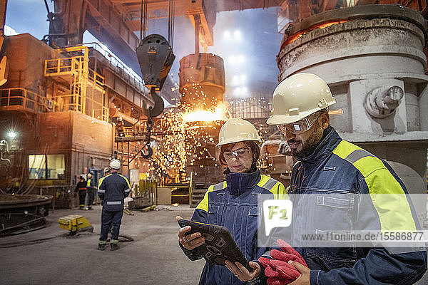 Male and female steelworkers using digital tablet during steel pour in steelworks