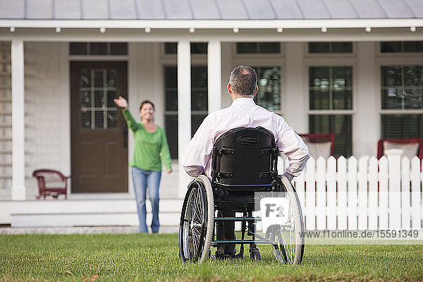 Frau winkt von ihrem Haus aus ihrem Mann im Rollstuhl zu  der eine Rückenmarksverletzung hat