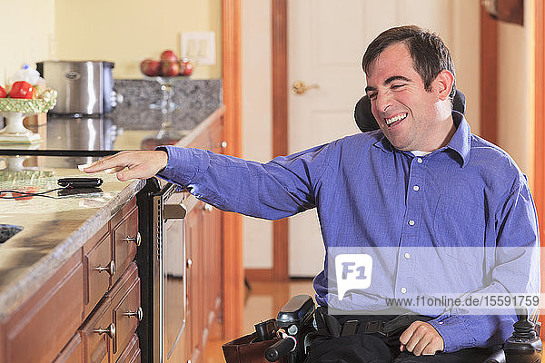 Mann mit Cerebralparese in seinem motorisierten Rollstuhl in seiner Küche greift nach seinem Handy