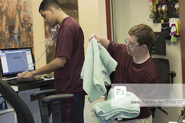 Young man with Down Syndrome preparing towels at college equipment dispensary for gym with supervisor using phone