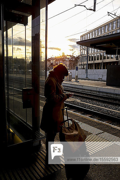 A woman stands with her suitcase on the platform of a train station beside the tracks and uses her smart phone; St. Gallen  St. Gallen  Switzerland