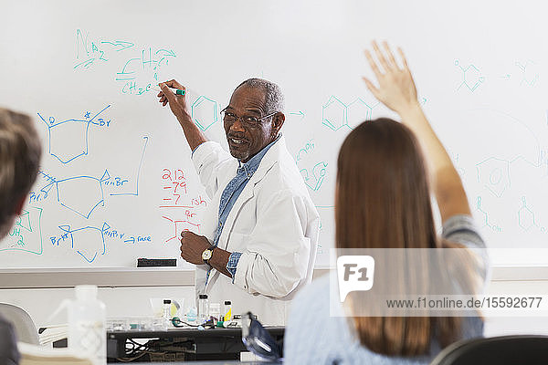 Student raising her hand in an engineering class while professor is at the white board writing about chemical bonds