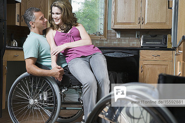 Eine mittelgroße Frau sitzt auf dem Schoß eines mittelgroßen Mannes im Rollstuhl und lächelt