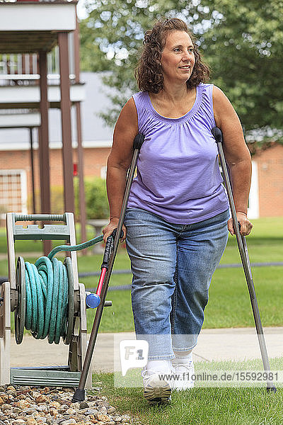 Frau mit Spina Bifida geht mit Krücken und zieht einen Gartenschlauch