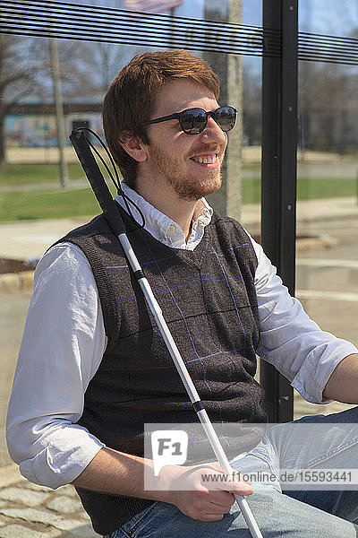 Junger blinder Mann mit Blindenstock wartet an einer Bushaltestelle
