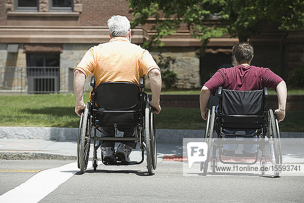 Rückansicht eines älteren Mannes mit Muskeldystrophie und einer jungen Frau  die in Rollstühlen eine Straße überqueren