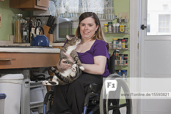 Frau mit Spina Bifida im Rollstuhl spielt mit einer Katze in der Küche