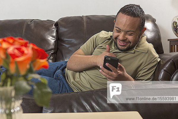 Glücklicher afroamerikanischer Mann mit Down-Syndrom  der zu Hause auf dem Sofa ein Mobiltelefon benutzt