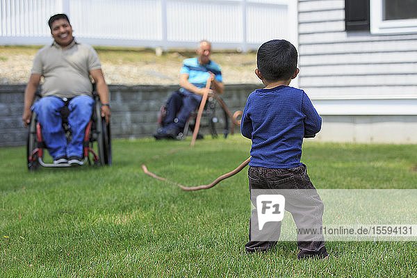 Junge  der mit einem Schlauch spielt  begleitet von Männern mit Rückenmarksverletzungen in Rollstühlen auf einer Wiese