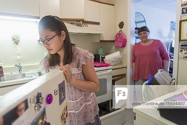 Lernbehindertes Teenager-Mädchen  das in den Kühlschrank schaut  mit ihrer Mutter im Hintergrund