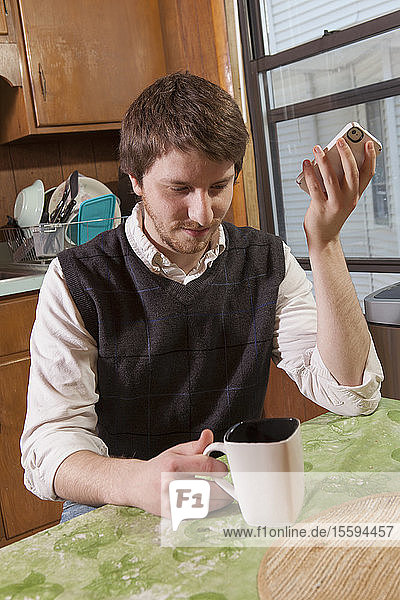 Blinder Mann in seiner Küche  der beim Kaffeetrinken Hilfsmittel auf seinem Mobiltelefon hört