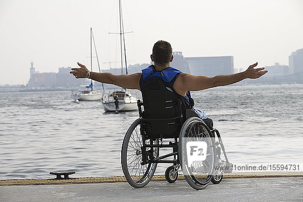 Rückansicht eines im Rollstuhl sitzenden Mannes mit ausgestrecktem Arm an einem Hafen