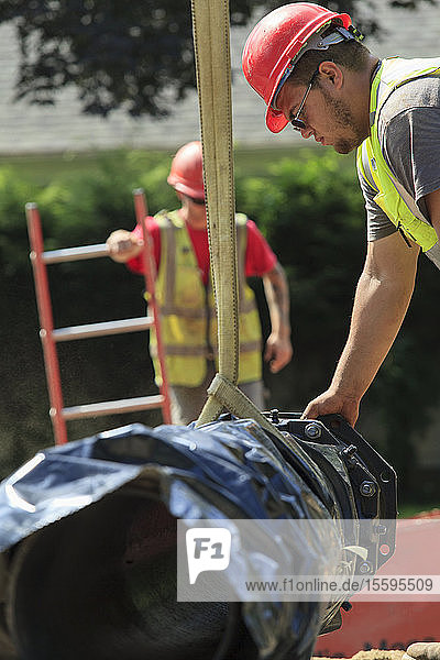 Bauarbeiter benutzt einen Gurt am Bagger  um einen Abschnitt der Wasserleitung zu führen
