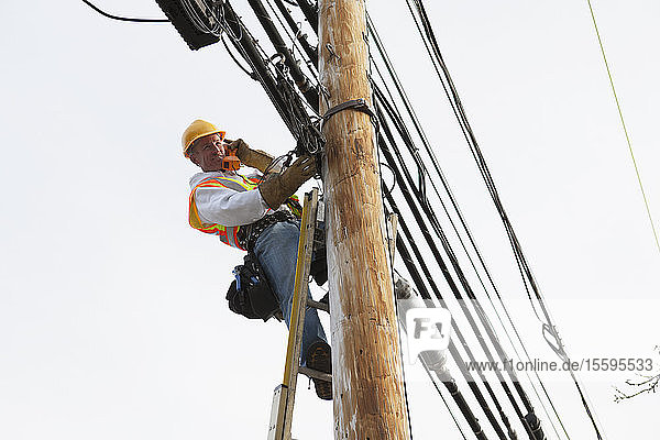 Ein Mitarbeiter der Kommunikationsbranche spricht am Telefon  während er neue Kabel an einem Strommast verlegt