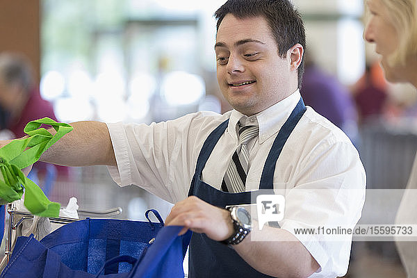 Mann mit Down-Syndrom arbeitet in einem Lebensmittelgeschäft und begrüßt einen Kunden beim Einpacken