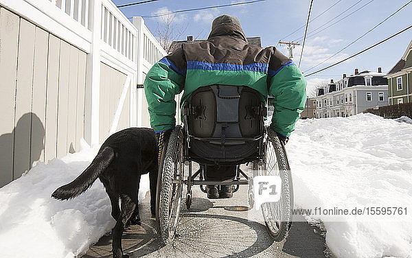 Frau mit Multipler Sklerose in einem Rollstuhl mit einem Diensthund auf einer verschneiten Straße
