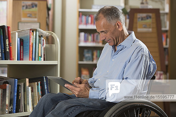 Mann mit einer Rückenmarksverletzung in einer Bibliothek beim Lesen eines Tablets