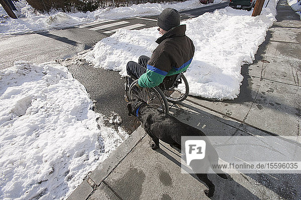 Eine Frau mit Multipler Sklerose im Rollstuhl mit einem Diensthund versucht  im Winter über einen Bordstein zu kommen