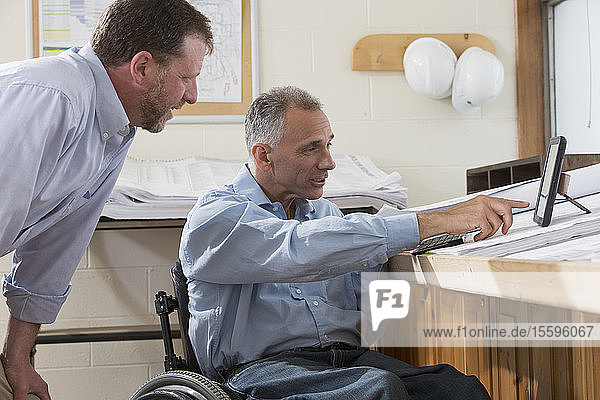 Zwei Projektingenieure nutzen ihr Tablet  um Pläne für die Baustelle zu prüfen  einer sitzt im Rollstuhl und hat eine Rückenmarksverletzung