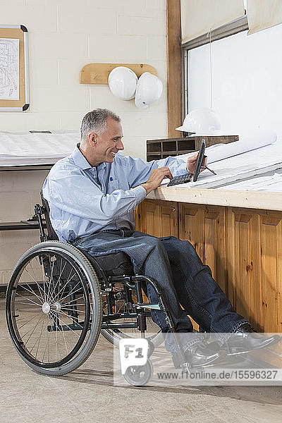 Projektingenieur  der im Rollstuhl sitzend mit einer Rückenmarksverletzung ein Tablet benutzt  um Pläne für die Baustelle zu prüfen