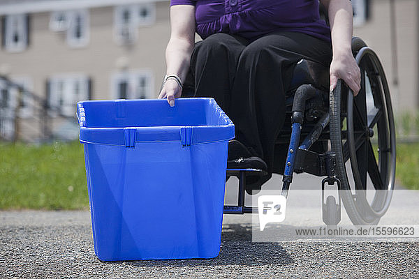 Frau mit Spina Bifida im Rollstuhl beim Abholen einer Recycling-Tonne an der Straße