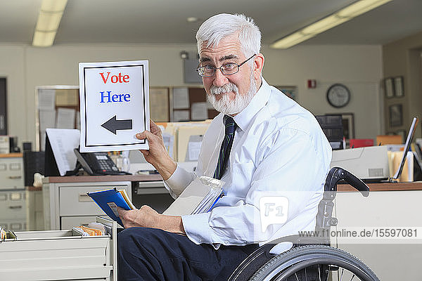 Ein Mann mit Muskeldystrophie im Rollstuhl arbeitet in seinem Büro und hält ein Schild mit der Aufschrift Vote Here hoch.