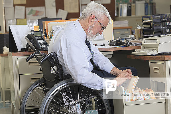 Mann mit Muskeldystrophie im Rollstuhl  der in der Schublade seines Büros Papierkram nachschlägt
