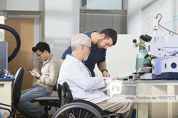 Professor mit Muskeldystrophie bei der Arbeit mit einem Ingenieurstudenten  der in einem Labor das Buch für den Röntgenfluoreszenzanalysator studiert