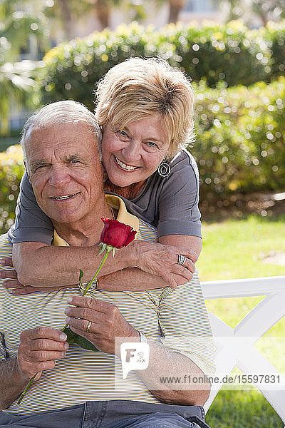 Portrait of a senior woman hugging a senior man from behind with a rose
