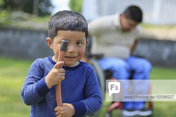 Portrait of Hispanic child holding hose and his father in wheelchair with Spinal Cord Injury in the background