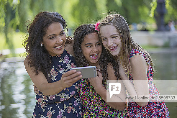 Glückliche hispanische Mutter mit zwei Teenager-Töchtern mit Zahnspange  die im Park Textnachrichten auf ihrem Handy lesen