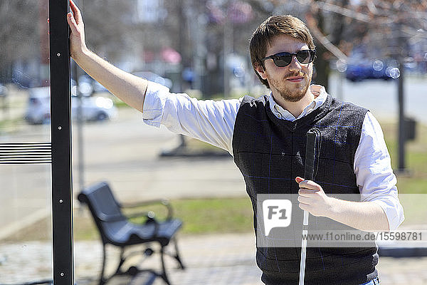 Junger blinder Mann mit Blindenstock wartet an einer Bushaltestelle