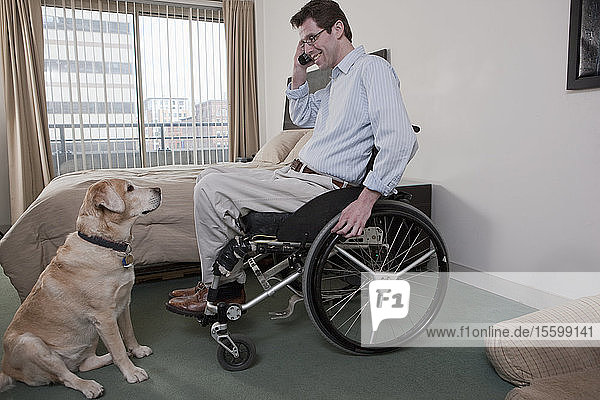 Ein Mann im Rollstuhl mit einer Rückenmarksverletzung spricht mit einem Diensthund über ein Mobiltelefon