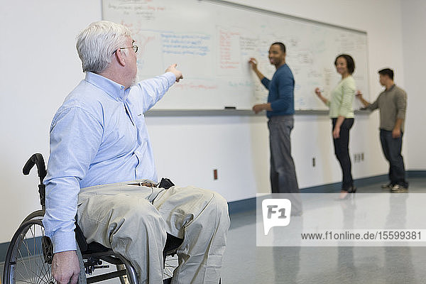 Universitätsprofessor mit Muskeldystrophie im Rollstuhl zeigt auf seine Studenten  die auf eine Tafel schreiben