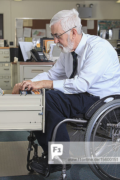 Mann mit Muskeldystrophie im Rollstuhl beim Abheften von Papieren in seiner Büroschublade