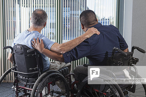 Zwei Männer mit Rückenmarksverletzungen in Rollstühlen  die durch ein Bürofenster hinausschauen und die Arme um sich legen