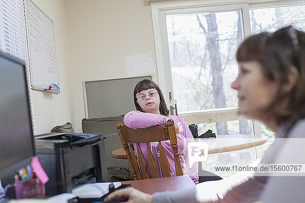 Mädchen mit Down-Syndrom  das seine Mutter bei der Arbeit am Computer beobachtet
