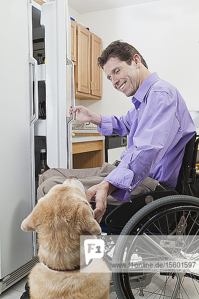 Mann im Rollstuhl mit Rückenmarksverletzung öffnet einen Kühlschrank und streichelt seinen Diensthund